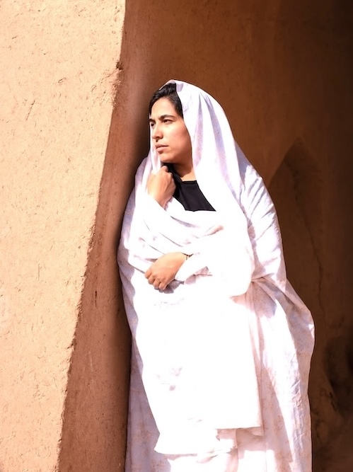 femme en haïek adossée à un mur de pisé brulé par le soleil de l'été.